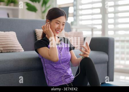Lächelnde asiatische Erwachsene Frau genießt persönliche Freizeit, um Audioinhalte über Smartphone und kabelgebundene Ohrhörer in gemütlicher Umgebung zu hören Stockfoto