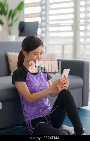 Glückliche asiatische Frau entspannt sich zu Hause und genießt Audiounterhaltung über Kopfhörer und Smartphone auf einer Matte in ihrem modernen Wohnbereich. Stockfoto