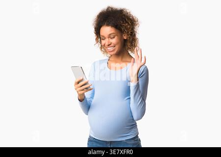 Lächelnde gemischte schwangere Frau stehend und mit Smartphone im Studio mit weißem Hintergrund Stockfoto
