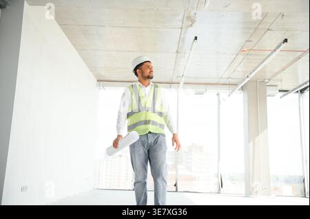 Indischer Ingenieur mit Sicherheitsweste und Schutzhelm mit Blaupausen beim Gehen und bei der Inspektion der Baustelle Stockfoto