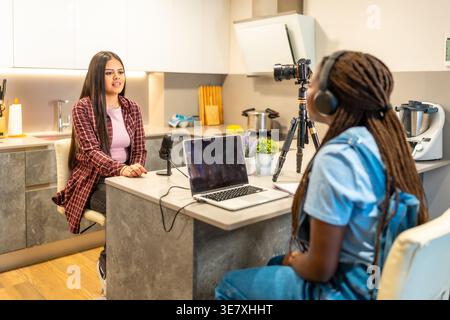 Hispanic und Black weibliche Podcaster, die Podcasts mit der Kamera aufnehmen, während sie zu Hause am Tisch sitzen Stockfoto