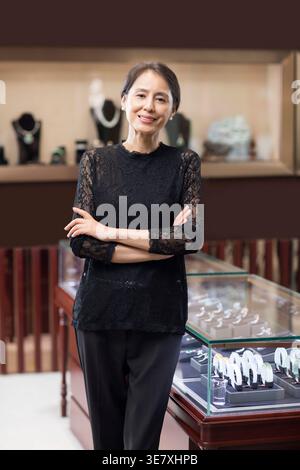Lächelnde chinesische Käuferin mittleren Alters in schwarzem Kleid, die mit überkreuzten Armen neben einem Glasvitrine im Juweliergeschäft steht Stockfoto