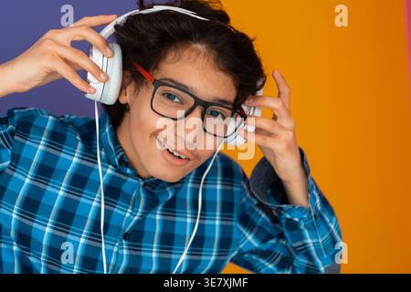 Lächelnder männlicher Teenager aus dem Nahen Osten in karierten langen Ärmeln und Brillen, der Musik über Kopfhörer lauscht, während er auf gelbem Hintergrund steht... Stockfoto