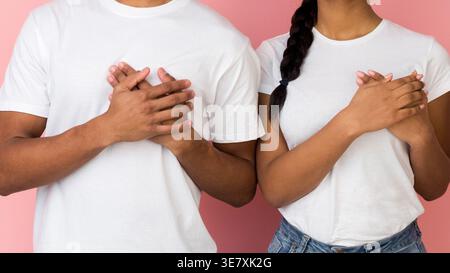 Schwarzes Paar hält die Hände auf der Brust zusammen, während es im Studio vor rosa Hintergrund steht Stockfoto