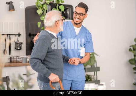 Lächelnder arabischer männlicher Betreuer in Brille, der einen älteren Mann im Stehen zu Hause ansieht Stockfoto