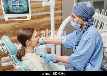 Gemischte Rassenzahnärztin mit Operationsmaske, die in der Klinik auf dem Zahnstuhl liegend eine kaukasierin mit Lätzchen belegt Stockfoto