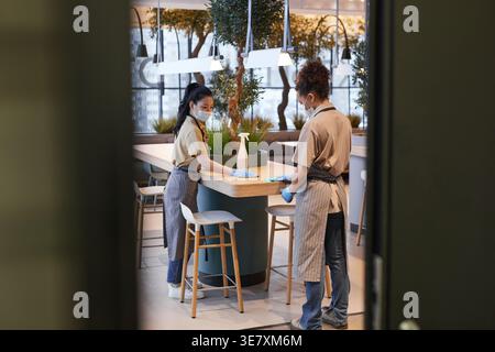Asiatische und gemischte Kellnerinnen mit Gesichtsmasken und Schürzen, die während der Pandemie im Café stehen Stockfoto