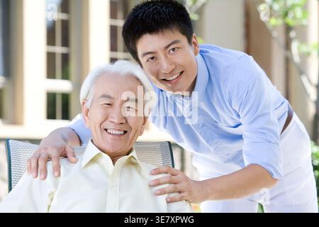Ein lächelnder chinesischer Mann sitzt tagsüber auf einem Stuhl und sein Sohn steht hinter ihm Stockfoto