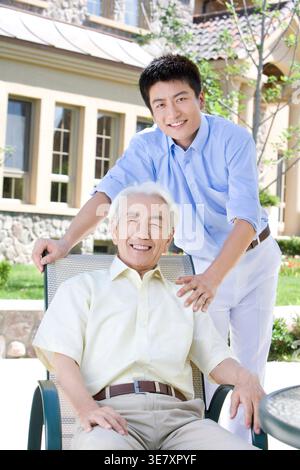 Ein lächelnder chinesischer Mann sitzt tagsüber auf einem Stuhl und sein Sohn steht hinter ihm Stockfoto