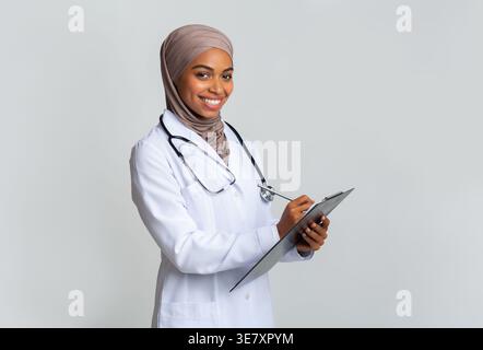 Schwarze muslimische Ärztin mit Stethoskop, die Hijab-Schrift auf der Zwischenablage trägt, während sie im Studio vor grauem Hintergrund steht Stockfoto