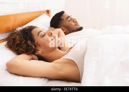 Schwarzes Paar schläft bequem mit Händen hinter dem Kopf, während es zu Hause unter weißer Decke auf dem Bett liegt Stockfoto