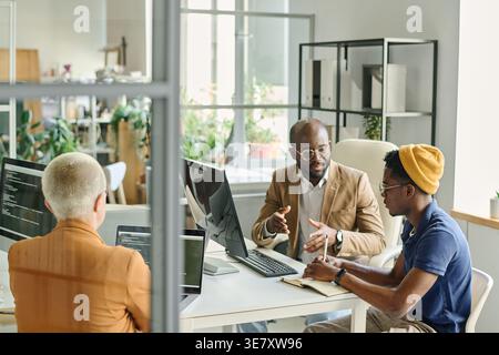 Serious Black, männliche Programmierer diskutieren über neues Projekt, während sie am Schreibtisch in der Nähe einer kaukasischen Kollegin sitzen, die im Büro kodiert Stockfoto