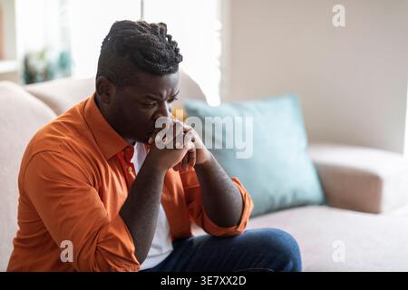 Ernster Schwarzer Mann, der den Mund auf gefesselte Hände lehnt, sitzt auf Sofa im Wohnzimmer zu Hause Stockfoto