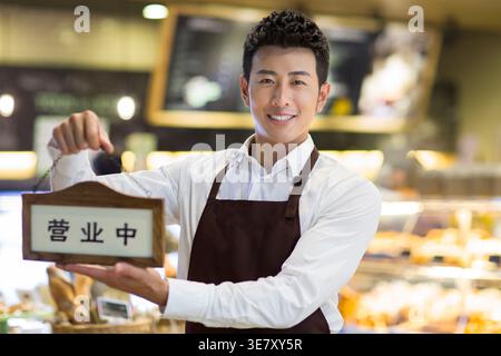 Ein lächelnder Chinese in brauner Schürze, der ein offenes Schild hält, während er in der Bäckerei steht Stockfoto