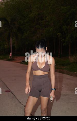 Eine junge thailändische Fitnessbegeisterte in einem braunen Neckholder-Top und Shorts mit hohem Bund steht nachts auf einem Outdoor-Pfad und überprüft nach einem W ihr Formular oder Telefon Stockfoto