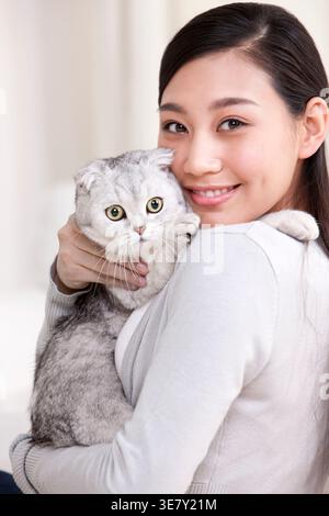 Lächelnde Chinesin, die schottische Faltkatze trägt und umarmt, während sie zu Hause auf dem Sofa sitzt Stockfoto