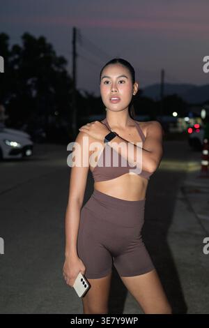 Eine junge Thai-Frau in brauner, gerippter Aktivkleidung berührt ihre Schulter, während sie ein Smartphone hält, während sie während eines abendlichen Workouts im Freien in Thailand trainiert, Representin Stockfoto