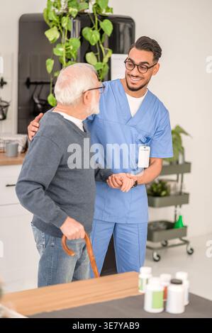 Lächelnder arabischer männlicher Betreuer in Brille, der einen älteren Mann im Stehen zu Hause ansieht Stockfoto