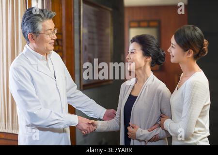 Lächelnder chinesischer Seniorarzt schüttelt nach der Untersuchung in der Klinik mit einer Patientin mittleren Alters mit ihrer Tochter die Hand Stockfoto