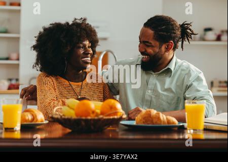 Fröhliches schwarzes Paar lacht und umarmt, während es am Tisch sitzt und zu Hause frühstückt Stockfoto