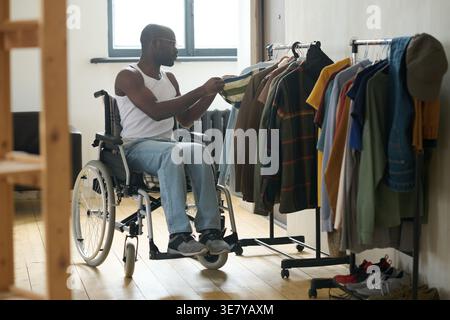 Behindertengerechter Schwarzer Mann, der im Rollstuhl sitzt und Kleidung zum Aufhängen zu Hause auswählt Stockfoto