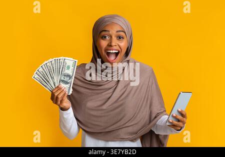 Schwarze muslimische Frau im Hijab, die Bargeld hält, während sie im Studio vor gelbem Hintergrund steht Stockfoto