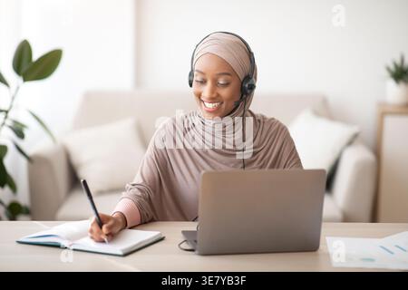 Schwarze muslimische Geschäftsfrau mit Headset, die Notizen vor dem Laptop am Tisch im Büro schreibt Stockfoto