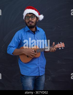 Lächelnder indischer Mann mit Santa Claus Hut mit Brille und blauen langen Ärmeln, während er Ukulele vor Tafel Hintergrund spielt Stockfoto