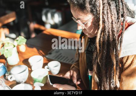 Hispanische Frau mit Dreadlocks, die eine Brille mit Smartphone trägt und tagsüber am Tisch mit Kaffeetassen im Café steht Stockfoto