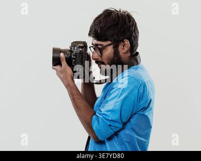 Lächelnder indischer Mann in blauen langen Ärmeln mit Brille, der Fotos mit einer DSLR-Kamera macht, während er im Studio vor weißem Hintergrund steht Stockfoto