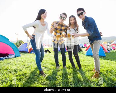 Lächelnde chinesische Freundinnen und Freundinnen, die beim Musikfestival zusammen auf dem Konzertgelände mit Zelten eine Gläser mit Bier anschlagen Stockfoto