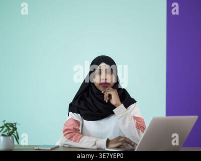 Ernsthafte schwarze muslimische Geschäftsfrau mit Hijab-Kinn auf der Hand, während sie mit Laptop im Büro am Tisch sitzt Stockfoto