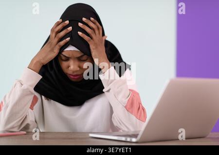 Gestresste schwarze muslimische Geschäftsfrau mit Händen auf dem Kopf und geschlossenen Augen, die im Büro mit Laptop und Notebook am Tisch sitzt Stockfoto