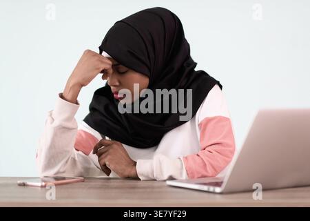 Gestresste schwarze muslimische Geschäftsfrau mit Hand auf Kopf und geschlossenen Augen, die im Büro mit Laptop und Smartphone am Tisch sitzt Stockfoto