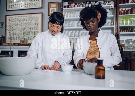 Japanische Apothekerin und schwarze Kollegin, die traditionelle Medizin verpackt, stehen in der Vintage-Apotheke Stockfoto