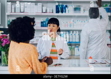 Japanische Apothekerin steht am Tresen und spricht mit schwarzer Kundin, als schwarzer Apotheke-Assistent Medikamente von der sie... Stockfoto