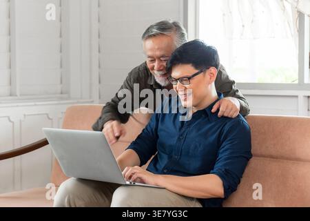 Asiatischer Seniorenvater, der hinter ihm steht und auf den Laptop blickt, mit einem Mann mittleren Alters, der zu Hause auf der Couch sitzt Stockfoto