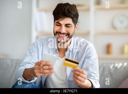 Lächelnder arabischer Mann mit Bart, der das Smartphone benutzt und die Kreditkarte hält, sitzt auf dem Sofa im Wohnzimmer zu Hause Stockfoto