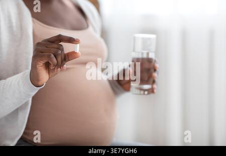 Schwarze schwangere Frau mit Vitaminpille Kapsel und Glas Wasser, während sie zu Hause auf dem Bett im Schlafzimmer sitzt Stockfoto