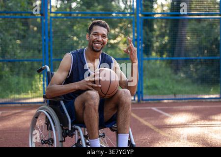 Schwarzer Mann mit Behinderung, der Basketball hält und ein OK-Schild zeigt, der tagsüber im Rollstuhl auf dem Basketballfeld sitzt Stockfoto