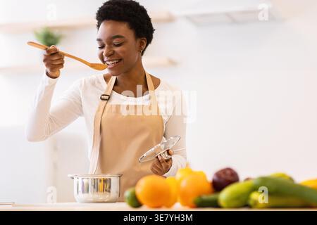 Schwarze Frau mit kurzen Haaren, die Schürze trägt, Kochen und Verkochen mit Holzlöffel, während sie zu Hause in der Küche steht Stockfoto