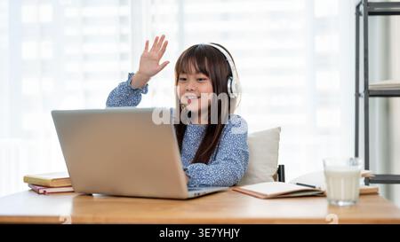 Asiatisches Mädchen mit Kopfhörern, die die Hand heben, während es online am Tisch sitzt und zu Hause Bücher trägt Stockfoto
