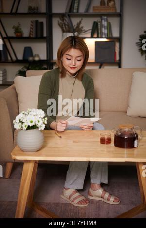 Lächelnde asiatische Frau, die einen Brief liest, während sie auf dem Sofa zu Hause Tee trinkt Stockfoto