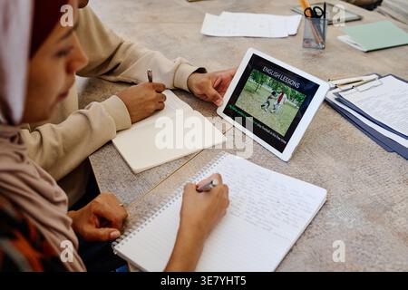 Hispanische muslimische weibliche und schwarze männliche Einwanderer sitzen am Tisch und schauen sich gemeinsam ein Lehrvideo auf einem Tablet an und machen sich Notizen während Englisch les... Stockfoto