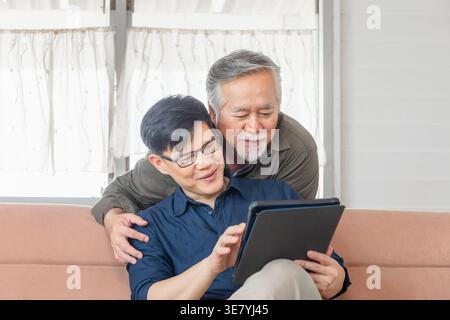 Asiatischer älterer Vater, der sich hinter seinem Sohn mittleren Alters mit einer Brille in der Hand eines Tablets auf der Couch zu Hause liegend Stockfoto