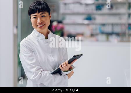 Fröhliche japanische Apothekerin mit Tablette im Arm, die in der Apotheke gegen den Zähler steht Stockfoto