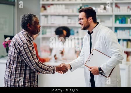 Senior Black man schüttelt die Hand mit einem männlichen kaukasier, der ein Klemmbrett hält, der in der Apotheke gegen den Tresen steht Stockfoto