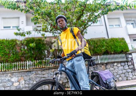 Schwarzer Mann mit Helm und Liefertasche, der tagsüber mit dem Fahrrad auf der Straße steht Stockfoto