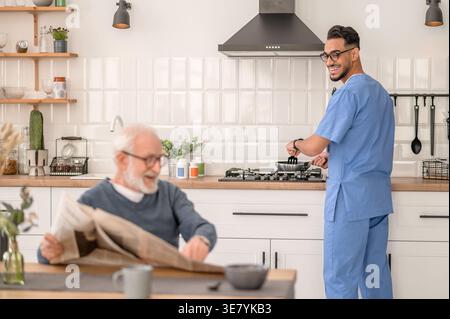 Lächelnder arabischer männlicher Pfleger in Brille kocht im Stehen, während er die Zeitung ansieht, wie ein älterer Mann zu Hause am Tisch sitzt Stockfoto