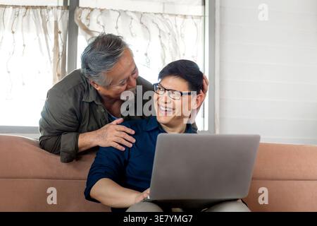 Asiatischer Mann mittleren Alters mit Laptop, der Spaß hat, wenn der ältere Vater hinter ihm steht, während er zu Hause auf der Couch sitzt Stockfoto
